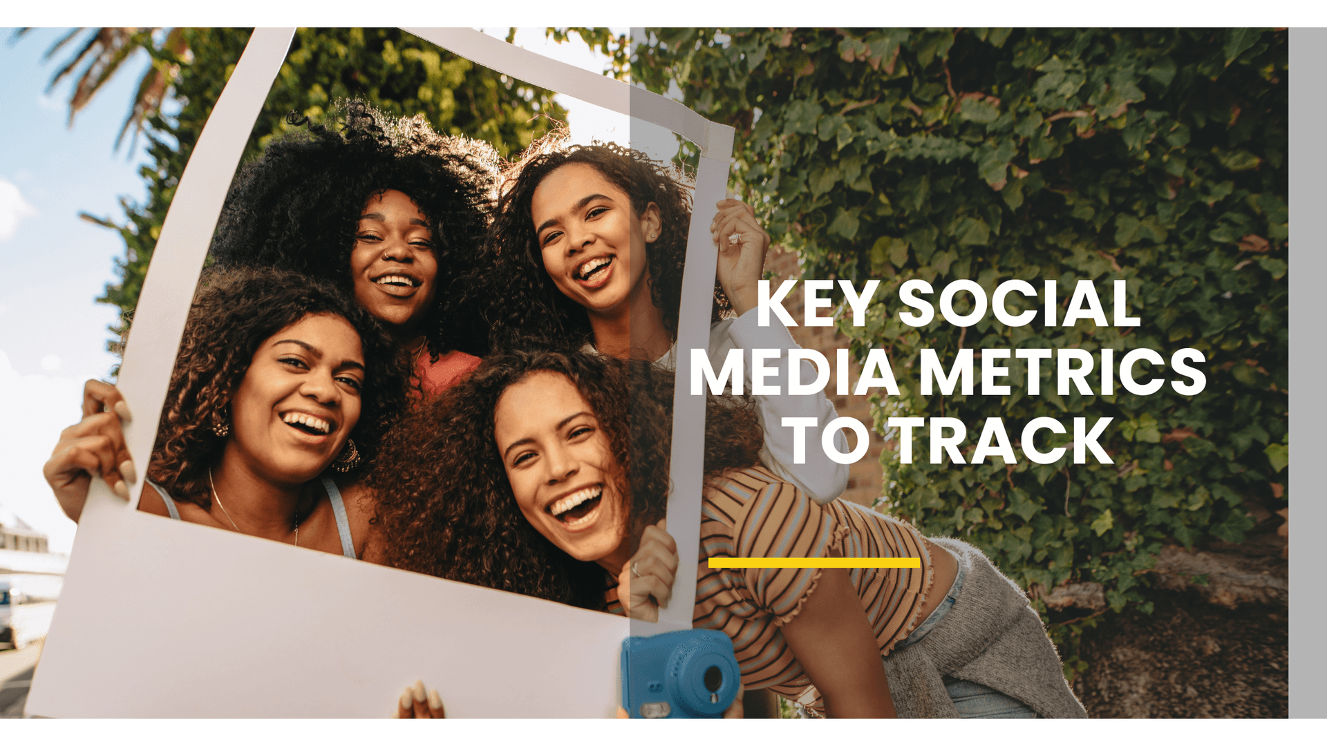 A group of four young women with curly hair are smiling and laughing outdoors, holding a large white photo frame prop in front of them. They are standing under a clear sky with palm trees in the background, giving a warm and joyful atmosphere.